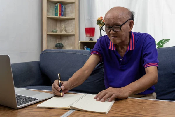 Asian elderly man holding pen write message note and computer laptop on table at sofa.
