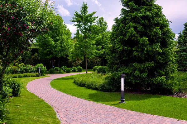 Güneşli bir yaz gününde yeşil çimlerle çevrili bahçeler, çalılar ve ağaçların arasında bahçeye taş döşemeler döşenmiş bir park yolunun fener demiri bahçe aydınlatması, hiç kimse.