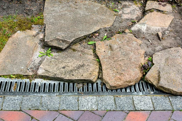 iron grating line of the drainage system of the pedestrian sidewalk made of stone tiles landscaping with natural rough stone of the park with engineering systems on side of the path, nobody.