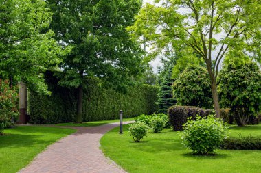Güneşli yaz gününde çimlerle çevrili bahçeler, çalılar ve ağaçların arasında, bahçesinde tuğla kiremitlerle döşenmiş hilal geçitli park parkı aydınlatması..