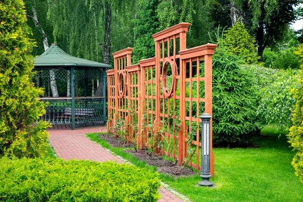 Vertical pergola made of wood in a rose garden with a stone tile walkway and ground lantern in backyard in a garden with plants with iron gazebo with a bench in the background.