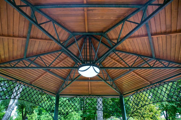 hanging lamp on the ceiling of the gazebo with an iron frame and wooden decorative inserts, a canopy in the backyard with an illuminator among plants close-up.