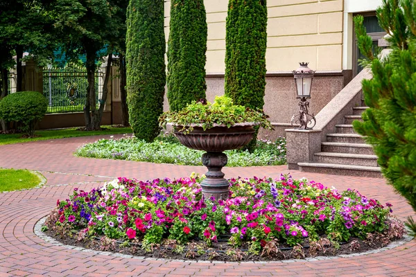 Stone flower bed fountain in the backyard with blooming petunias with a brick tile pavement in front of a granite building and an iron lantern street lighting, nobody.