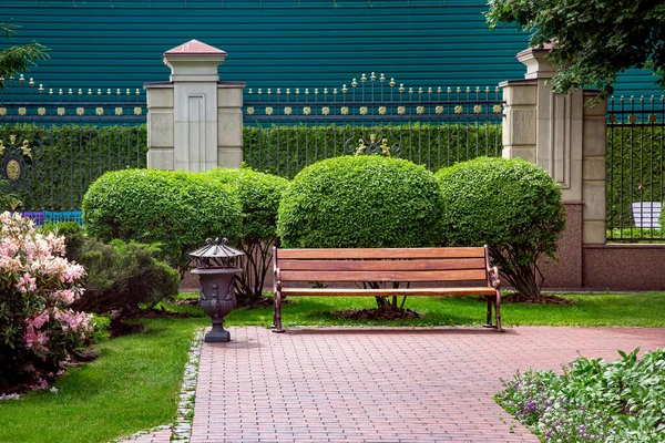 緑の植物や金属のピークフェンスと裏庭の庭の石のタイルに鉄くずビンと木製のベンチ 誰も座っていない空の座席と公園の春の風景 ロイヤリティフリーのストック写真