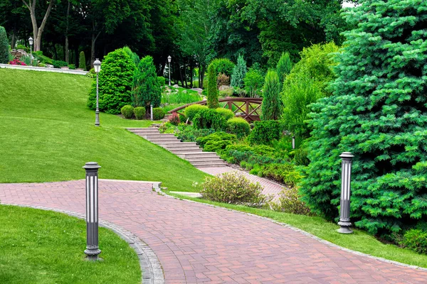 公园弧形道路上的铁灯地面和柱子花园照明 石砖铺成 园内植物 常绿灌木和松树中间有排水格栅 山水清澈 — 图库照片