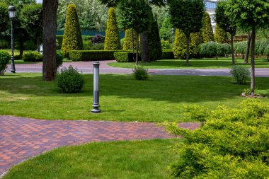 iron retro lanterns of street lighting ground and pillar in park with stone tile pedestrian walkway and green lawns with trees and evergreen arborvitae hedge in scenic parkland on summer day, nobody.