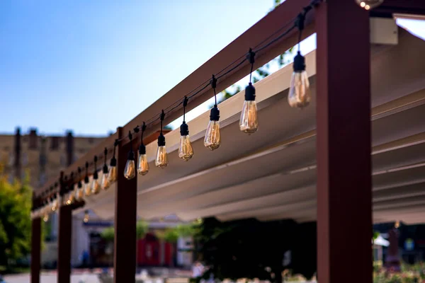 城市街道咖啡店中的纺织品雨篷木制框架花环 花环串串着复古的爱迪生灯泡 在夏日的近景中闪烁着温暖的光芒 没有人 — 图库照片