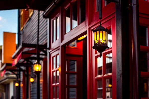hanging iron retro lanterns with warm glow on facade of decorative style wooden building with red entrance door frame with glass. Architecture of evening lighting of city streets decorative light.