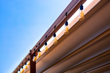 Arka bahçedeki tekstil tenteleri ahşap çerçeve çardağı retro-Edison çelenklerinden oluşan çelenkler terasta sıcak ışıklarla parıldıyor, yaz günü mavi gökyüzü ile yakın plan ışık dekoru, hiç kimse.