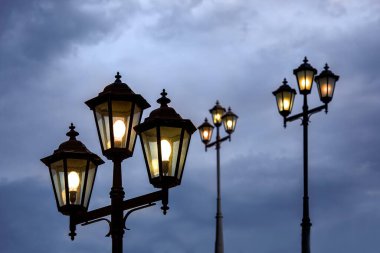 Şeffaf cam paneller ve elektrik ampulü ile üçlü fener baş, demir sokak aydınlatma akşam parlayan mavi bulutlu gökyüzü, retro tarzı lamba fikstürü karşı.