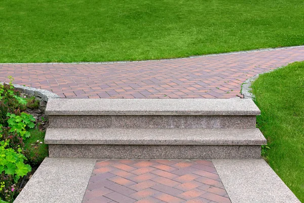 Yeşil çimenli bir araziyle çevrili taş kiremitlerden oluşan bir kaldırım boyunca uzanan yamaçta granit adımlar, bir şehir parkının peyzajına yakın çekim, hiç kimse.