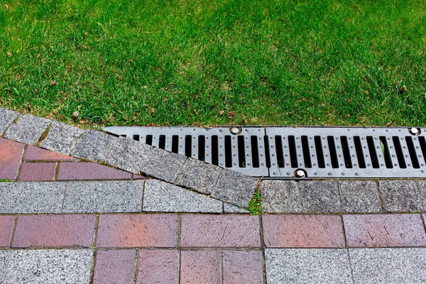drainage grate bolted to storm drain at corner of pavement walkway path made of stone brick tiles red and gray color with pattern in backyard with grass close-up of engineering structures of garden.