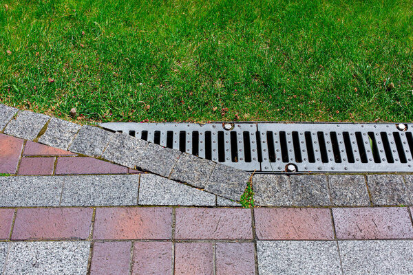 решетка дренажной решетки, прикрученная к ливневой канализации в углу тротуара из камня кирпичной плитки красного и серого цвета с узором на заднем дворе с травой крупным планом инженерных сооружений сада.