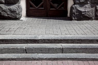 granite steps on a pedestrian pavement made of stone tiles near a building with entrance a wooden front door and a facade lined with rough stone on a sunny day, close-up, nobody.