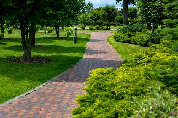Bahçenin arka bahçesinde tuğladan taş kiremitlerle döşenmiş park yolu bitkilerin, yemyeşil çalıların ve ağaçların arasında güneşli bir günde yeşil çimlerle çevrili, fener demiri parkında ışıklandırılmış, hiç kimse.