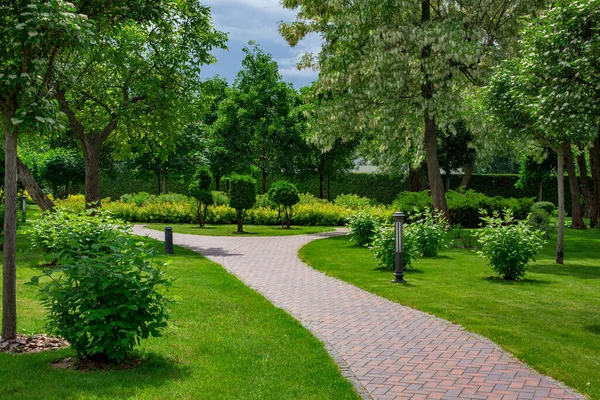 lantern iron ground garden lighting of curved park path paved with stone tiles in backyard among plants with mulching, bushes and trees surrounded by a green lawn on a sunny spring day, nobody.