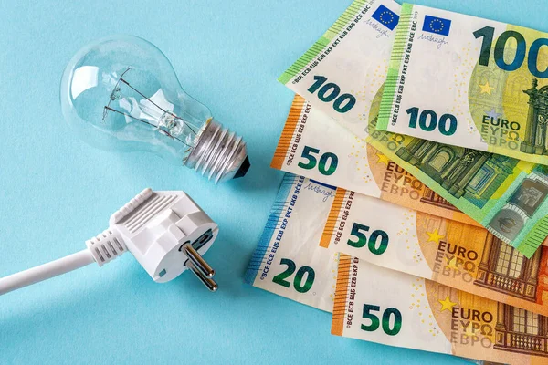 Beyaz elektrik prizi, ampul ve avroluk banknotlar mavi arka planda. Konut ve işyeri kullanıcıları için elektrik maliyetlerinin artırılması, pahalı enerji faturası ve elektrik fiyatları kavramlarındaki artış. 