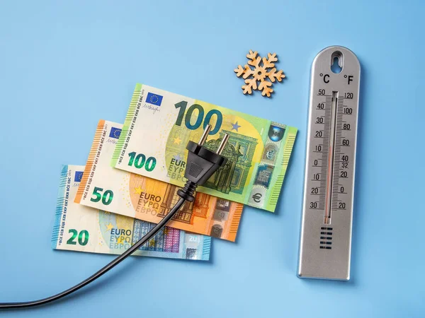 Siyah elektrik prizi 20, 50 ve 100 avronun üzerinde banknotlar, kar tanesi ve mavi zemin üzerinde hava termometresi. Soğuk kış mevsiminde elektrik fiyatlarında artış, elektrik kavramının maliyetinin artması. 