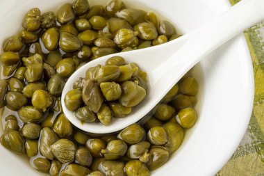 Kapta turşulu kapari dolu beyaz porselen kaşık. Kapari çalılarının marine edilmiş tomurcukları. Akdeniz mutfağı malzemesi. Organik baharatlar ve baharatlar. Makro atış. Üst görünüm.