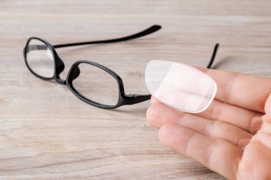 El, gözlüğün kırık kenarından dolayı lensin düştüğünü gösteriyor. Kahverengi bir masanın üzerinde kırık mor göz telleri olan gözlük. Okuma ve bilgisayar işleri için görüş düzeltme gözlüklerinin kırılması. Ön görünüm.