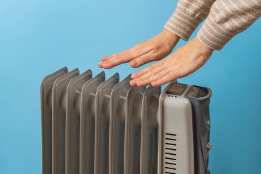 Kadın ellerini sıcak yağın yanında ısıtıyor. Mavi arka planda elektrikli ısıtıcı var. 9 yüzgeçli ısıtıcı kullanıyor. Soğuk mevsimde ısıtmak için taşınabilir ev aletleri. Ön görünüm.