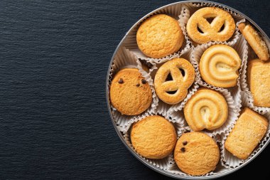 Açık bir teneke kutuda, siyah levha üzerinde gevrek bisküviler. Kahvaltıda lezzetli Danimarka kurabiyeleri var. Pişmiş pasta, tatlı yemek ve kalori kavramları. Uzayı kopyala Üst görünüm.