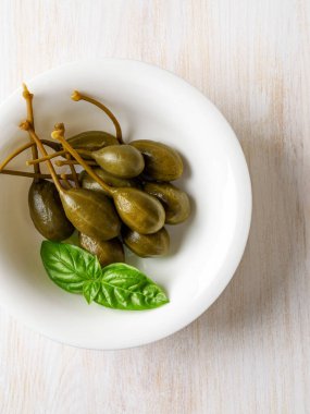 Kırsal bir masada beyaz bir kasede turşu ve taze fesleğen yaprağı. Tabakta marine edilmiş kapari üzümü. Baharat ve garnitür olarak konserve yaban mersini. Akdeniz mutfağı malzemesi. Üst görünüm.