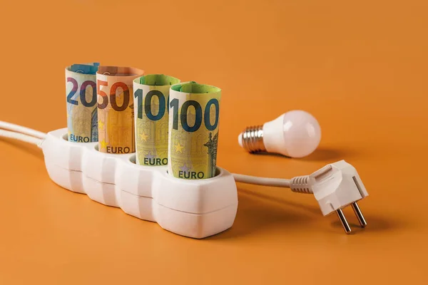 Rising energy costs concept. Euro money banknotes plugged into a white power strip, electrical plug and light bulb over orange background. High energy costs in Europe. Electricity bill payment. Front view.