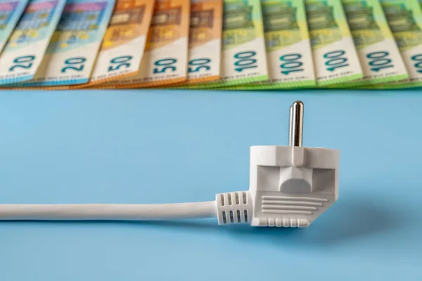 Rising power bills. Electricity price spike concept. White electrical plug in front of euro money banknotes row against blue background. High energy costs in Europe. Electricity cost. Front view.