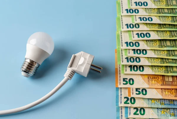 Rising energy prices. White electrical plug and light bulb at euro money banknotes over blue background. European power prices, electricity price spike, rising power bills concepts. Top view.