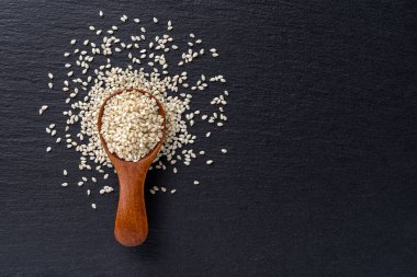Raw sesame seeds in a wooden spoon over black slate background. Sesamum indicum for healthy eating and boosting immunity diet. Organic benne grains as cuisine ingredient concept. Copy space. Top view.