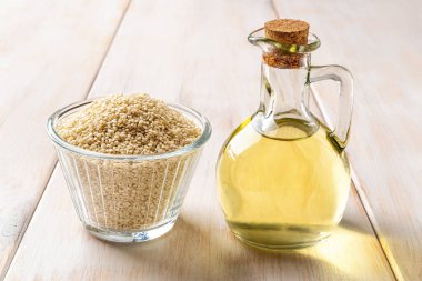 Fresh sesame oil in a glass jug and bowl of raw seeds on a wooden table. Sesamum indicum for oil production. White til for healthy food and boosting immunity diet. Asian cuisine ingredient. Front view.