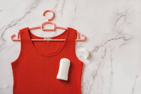 Red tank top on a hanger and solid antiperspirant over marble background. Deodorant stick on a linen jersey tank shirt. Prevent body odour, body care, toiletries concept. Copy space. Top view.