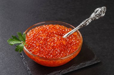 Bowl of chum salmon caviar on a black slate background. Trout red caviar and vintage spoon in a glass bowl close-up. Seafood fish delicacy of salted salmon roe. Gourmet food concept. Top view.