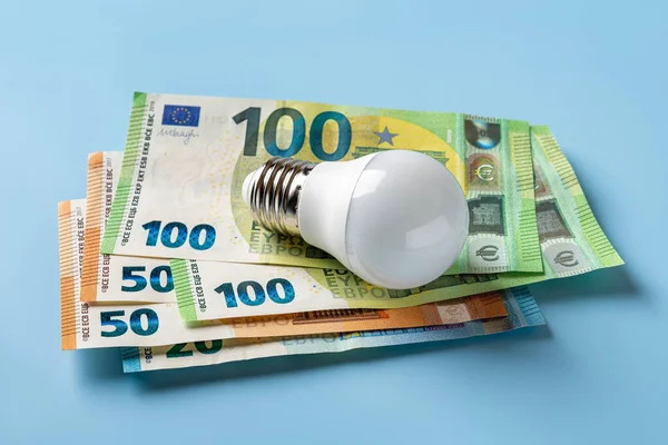 Light bulb on 50 and 100 euro currency banknotes over blue background. High energy costs concept. Power price rising in Europe. Soaring of electricity bills. Energy price spike. Top view.