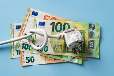 Knotted power cable with electric plug on 50 and 100 euro banknotes over blue background. Concept of rising energy costs in Europe. Electric bill increases. Power price spike. Top view.