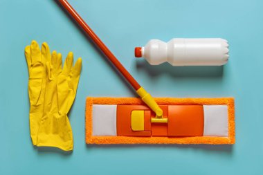 Flat mop, floor cleaner and rubber gloves over blue background. Orange microfiber mop with handle, white bottle of liquid cleaner, yellow rubber gloves flat lay. Mopping floors, housework. Top view.