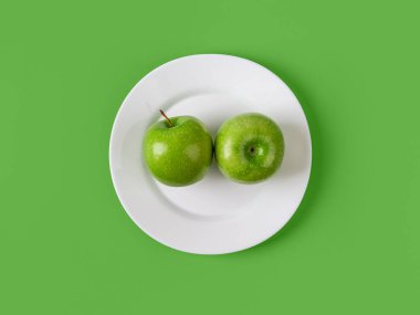 Yeşil arka planda beyaz bir tabakta iki taze yeşil elma. Zayıflamak ve kilo vermek için çiğ organik meyveler. Vitamin vejetaryen atıştırmalığı. Antioksidan sağlıklı beslenme. Üst görünüm.