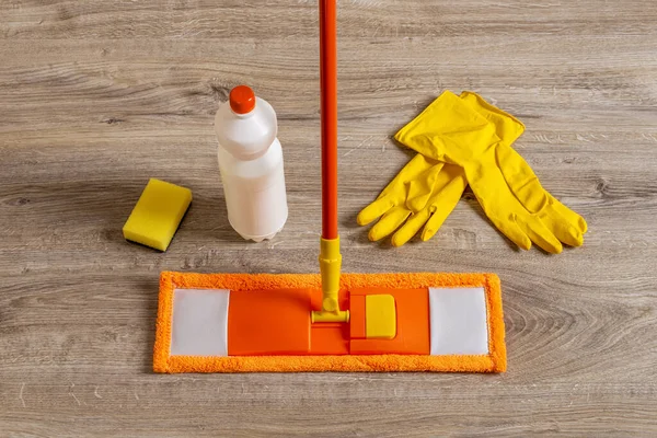 Flat mop and rubber gloves on the floor. Orange microfiber mop, yellow gloves, sponge and liquid cleaner bottle closeup. Mopping floors, homework cleaning routine, housekeeping concept. Top view.