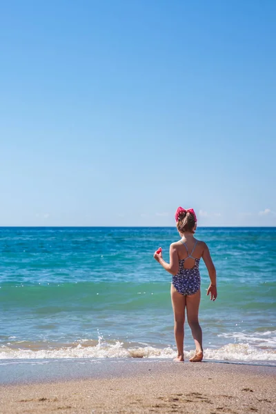 La chica está de pie con su espalda en la playa contra el telón de fondo de una hermosa ola Imágenes De Stock Sin Royalties Gratis