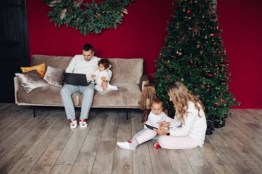 Baba ve oğul bir kanepede oturuyor, anne ve kız noel ağacının yanında oynuyorlar.