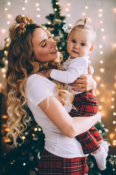 Atractiva madre joven sostiene a un bebé de rodillas en el ambiente navideño —  Fotos de Stock