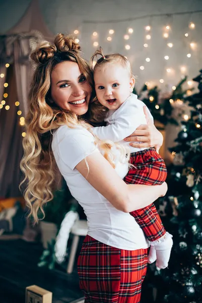 Hermosa joven madre sostiene a su bebé cerca del árbol de Navidad —  Fotos de Stock