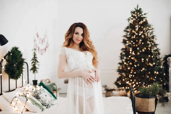 Femeia gravidă fermecătoare pozează pentru aparatul foto în rochie albă lângă pomul de Crăciun cu un salon de lumini — Fotografie, imagine de stoc