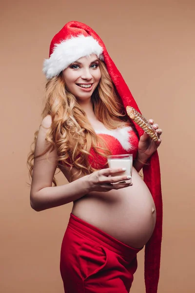 Tânără frumoasă femeie însărcinată în costum de Moș Crăciun mănâncă turtă dulce Tula cu lapte. — Fotografie, imagine de stoc