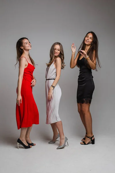 Encantadoras amigas jóvenes positivas con elegantes vestidos de noche femeninos posando sobre fondo de estudio gris — Foto de Stock