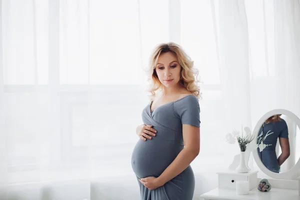 Hinreißende schwangere Frau im langen grauen Kleid, die ihren Bauch verstaut. — Stockfoto