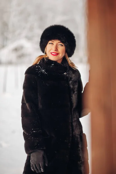 Seorang wanita muda yang cantik dengan topi bulu hitam dan mantel bulu, potret. Seorang gadis Rusia yang lucu tersenyum dan melihat ke dalam bingkai, bibir merah, riasan cerah. — Stok Foto