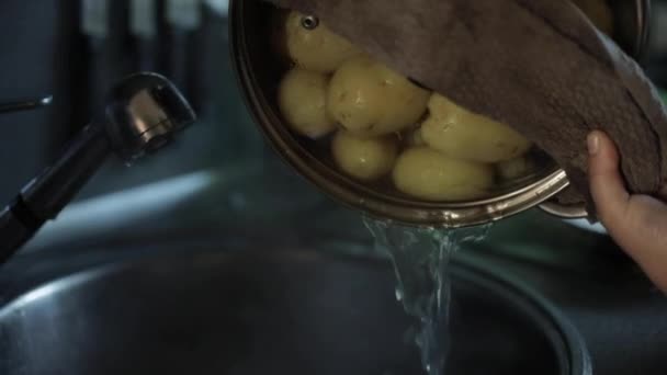 En hemmafru dränerar vatten från en gryta från en kokt potatis i diskhon. — Stockvideo