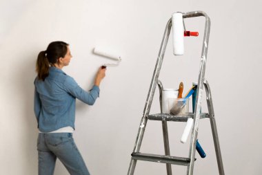 Boya fırçaları, kovalar ve aletlerle dolu bir merdiven. Arka planda duvar boyayan bir kadın. Ev geliştirme.